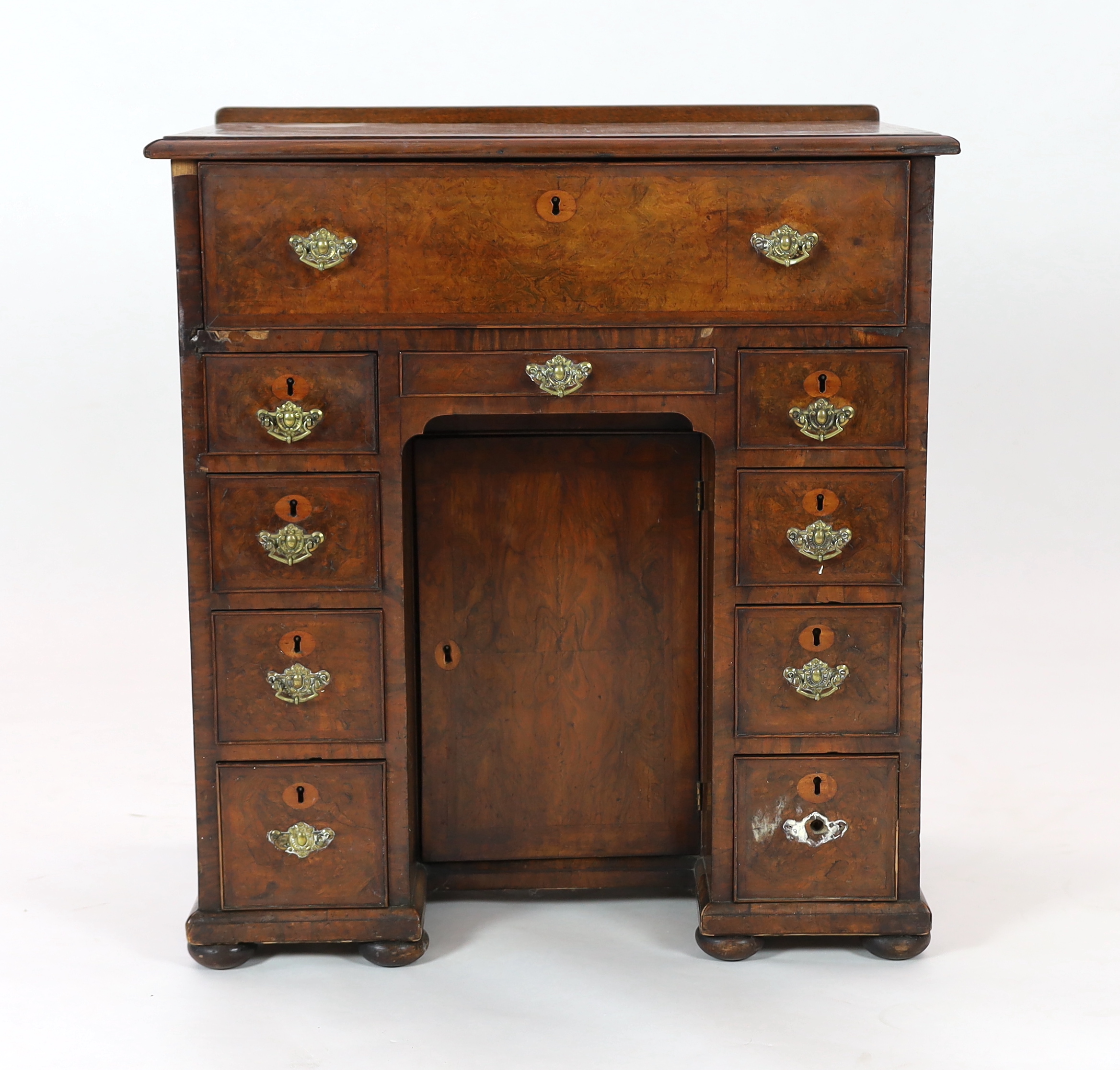 An early 18th century walnut kneehole secretaire desk, with quarter veneered top and fall front drawer opening to reveal pigeon holes and drawers, over eight further drawers surrounding a central recessed cupboard, on pa